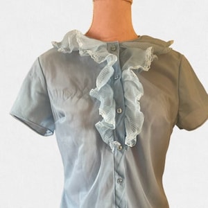 Könnte beinhalten: Eine hellblaue Bluse mit einem großen Rüschenkragen. Der Kragen ist aus einem durchsichtigen Stoff und hat einen zarten, femininen Look.