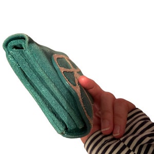 May include: A teal green leather wallet with a silver peace sign design.