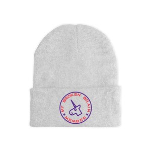 May include: A light gray beanie with a circular emblem. The emblem features a purple unicorn head and the words "MY BROKEN BRAIN MEMBER" in a circular design. The beanie is made of a soft, knitted material.