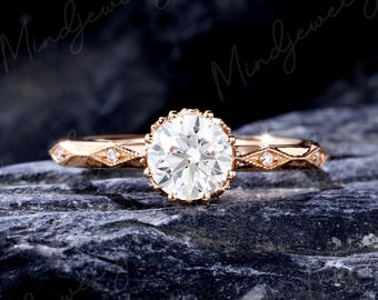 Könnte beinhalten: Verlobungsring aus Roségold mit einem großen, runden, klaren Edelstein. Der Ring hat ein detailliertes Band mit kleinen Diamantakzenten und eine dekorative Fassung um den Mittelstein. Der Ring wird auf einer dunklen, strukturierten Oberfläche präsentiert.