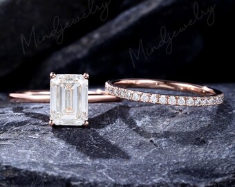 Op de afbeelding: Twee roségouden ringen. Eén ring heeft een grote, rechthoekige, heldere edelsteen. De tweede ring is een dunne band met kleine, heldere edelstenen. Beide ringen worden gepresenteerd op een donkere, getextureerde ondergrond.