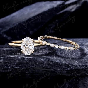 Puede incluir: Un anillo de compromiso de oro con un diamante de talla ovalada y una alianza de diamantes a juego. Los anillos se presentan sobre un fondo oscuro y texturizado. El diamante ovalado es el punto focal, con una banda delicada. La segunda banda está adornada con diamantes más pequeños.