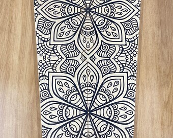 May include: A flat, tapered wooden board featuring a black ink mandala pattern printed on its surface. The design consists of two large, symmetrical floral-like geometric motifs with intricate petal shapes, curved lines, and dot details.