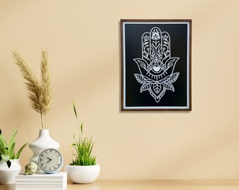 May include: A black and white framed print of a hamsa hand with an eye in the center, surrounded by floral designs. The print is hanging on a beige wall.