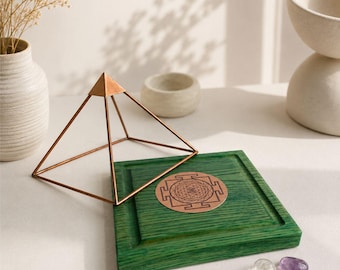 May include: A copper-colored wire pyramid frame positioned over a square green wooden base featuring a circular copper-colored Sri Yantra symbol inlay. Two small polished stones, one clear and one purple, rest near the base of the wooden square.