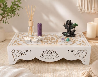 May include: A rectangular white wooden altar table featuring intricate gold-coloured floral and lotus carvings on the top and front panels. The table holds a glass reed diffuser, a purple pillar candle, three tea lights, and a dark stone incense burner.