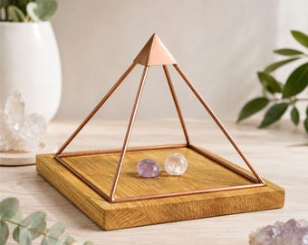 May include: A copper-colored metal pyramid frame sits on a square wooden base. Inside the frame, two small, smooth, polished stones are placed on the wood surface: one is a translucent light purple amethyst and the other is a clear quartz sphere.