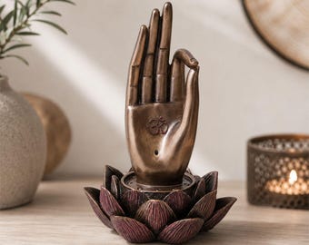 May include: A bronze-colored sculpture of an upright hand in a mudra gesture, featuring an Om symbol embossed on the palm. The hand is positioned within a dark, textured lotus flower base with layered petals, creating a decorative tabletop ornament.