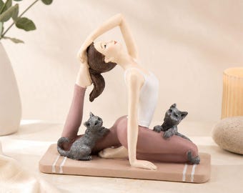 May include: A resin figurine of a person with brown hair in a ponytail, wearing a white tank top and mauve leggings, performing a yoga pose on a rectangular mat. Two small gray kittens are positioned near the person on the mat.