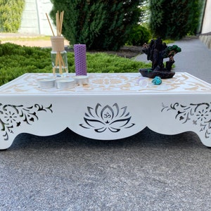Op de afbeelding: Een witte houten altaartafel met een lotusbloemdesign en het Om-symbool uitgesneden aan de voorkant. De tafel heeft een gouden bloemenpatroon op het bovenoppervlak. Kaarsen, wierook en kristallen worden op de tafel getoond.