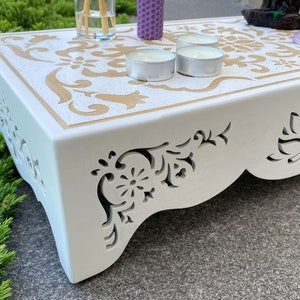 Op de afbeelding: Een witte houten altaartafel met een gouden bloemenontwerp op het bovenoppervlak. De zijkanten hebben ingewikkelde uitgesneden patronen. De tafel bevat kaarsen en andere decoratieve items, waardoor een serene en spirituele sfeer ontstaat.