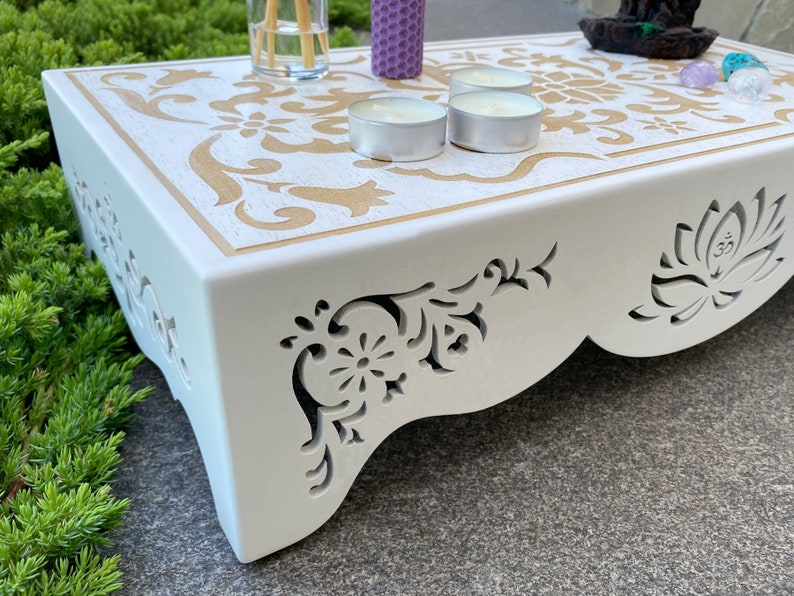 Op de afbeelding: Een witte houten altaartafel met een gouden bloemenontwerp op het bovenoppervlak. De zijkanten hebben ingewikkelde uitgesneden patronen. De tafel bevat kaarsen en andere decoratieve items, waardoor een serene en spirituele sfeer ontstaat.