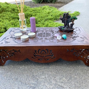 May include: A wooden altar with intricate cutouts, featuring a purple candle, a clear glass bottle with reeds, and a small ceramic incense burner with a tree on top. There are also three white tea lights and two small pink and turquoise stones on the altar.