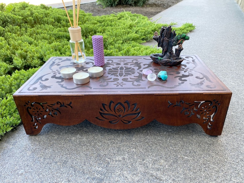 May include: A wooden altar with intricate cutouts, featuring a purple candle, a clear glass bottle with reeds, and a small ceramic incense burner with a tree on top. There are also three white tea lights and two small pink and turquoise stones on the altar.