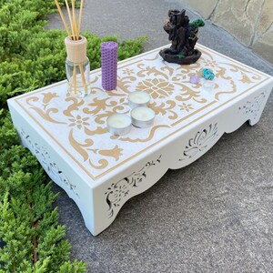 Op de afbeelding: Een witte decoratieve houten tafel met een gouden bloemenontwerp. De tafel heeft uitgesneden details en bevat kaarsen, kristallen en een wierookbrander. Een rietverspreider en een paarse kaars staan ook op de tafel.