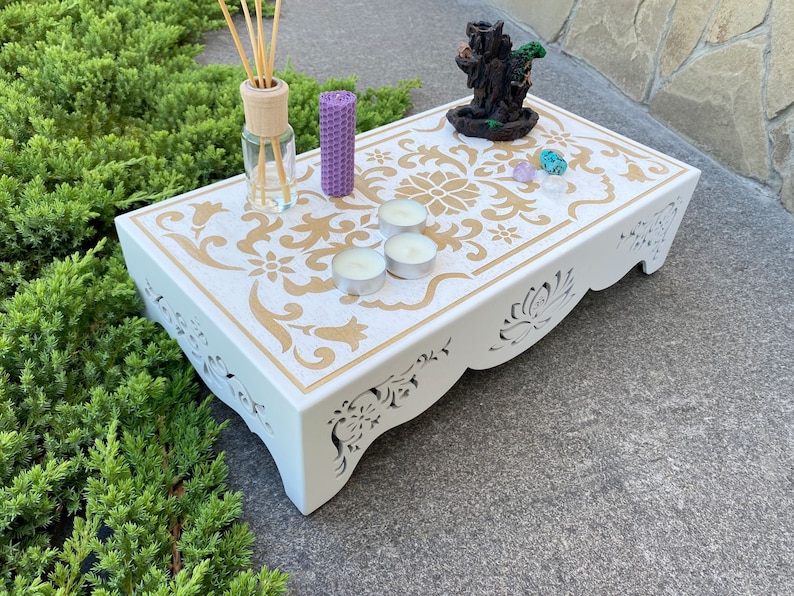 Op de afbeelding: Een witte decoratieve houten tafel met een gouden bloemenontwerp. De tafel heeft uitgesneden details en bevat kaarsen, kristallen en een wierookbrander. Een rietverspreider en een paarse kaars staan ook op de tafel.