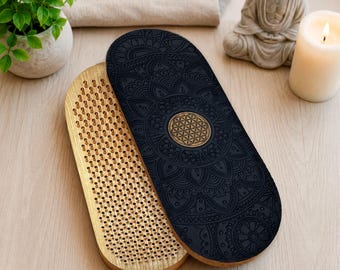 May include: An oval wooden sadhu board with a natural light wood finish on one side featuring a dense grid of metal nails. The other side is a dark black surface with an intricate mandala pattern and a central gold-toned flower of life symbol.