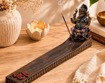 May include: A dark brown wooden incense burner featuring a sculpted Ganesha figure at one end and a red Om symbol at the other. The rectangular base has a decorative border and holds a single burning incense stick emitting a thin trail of smoke.