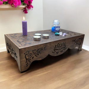 May include: A dark brown wooden altar table with carved floral and lotus designs. The table holds a purple candle, three tea lights, a jar of blue-coloured liquid, and small crystals. The table is on a wooden floor.