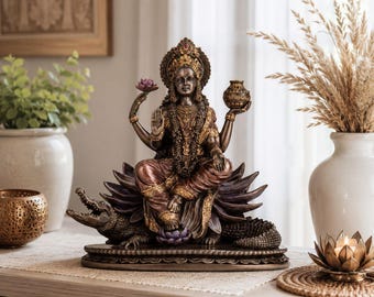 May include: A bronze-toned statue of a four-armed deity seated on a lotus flower atop a crocodile. The figure wears ornate jewelry and a crown, holding a lotus blossom in one hand and a small pot in the other. The sculpture features intricate detailing.