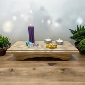 Puede incluir: Un altar de madera con una vela morada, tres velas de té blancas y varios cristales. El altar está sobre una mesa de madera.
