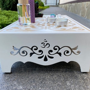Op de afbeelding: Een witte houten meditatietafel met een decoratief goudkleurig patroon en uitgesneden ontwerpen aan de zijkanten. De tafel bevat een diffuser, kaarsen en kristallen, wat een serene sfeer cre&euml;ert. Ontworpen voor meditatie en spirituele praktijken.