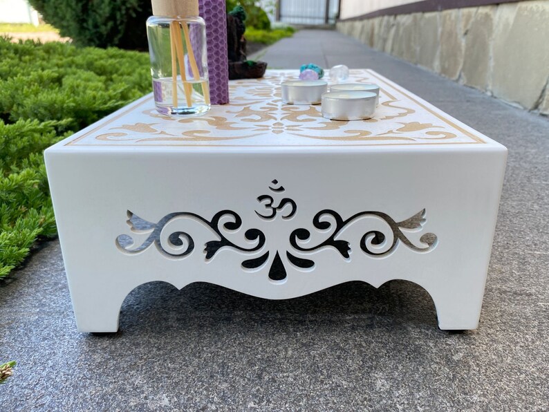 Op de afbeelding: Een witte houten meditatietafel met een decoratief goudkleurig patroon en uitgesneden ontwerpen aan de zijkanten. De tafel bevat een diffuser, kaarsen en kristallen, wat een serene sfeer cre&euml;ert. Ontworpen voor meditatie en spirituele praktijken.