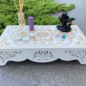 Op de afbeelding: Witte houten altaartafel met gouden bloemenontwerp en uitgesneden lotusbloem. Inclusief kaarsen, een geurverspreider, een paarse kaars en een kleine waterval wierookbrander. De tafel is ongeveer 46 cm lang.
