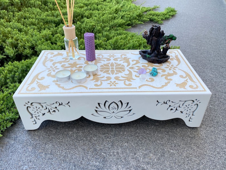 Op de afbeelding: Witte houten altaartafel met gouden bloemenontwerp en uitgesneden lotusbloem. Inclusief kaarsen, een geurverspreider, een paarse kaars en een kleine waterval wierookbrander. De tafel is ongeveer 46 cm lang.