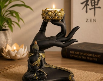 May include: A black and gold resin sculpture featuring a seated Buddha figure at the base and a large hand reaching upwards to hold a lotus-shaped candle holder. The sculpture includes intricate gold detailing on the Buddha's robe and the base.
