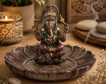 May include: A metallic silver-toned Ganesha figurine seated on a dark, textured lotus-shaped incense burner. The deity features green beaded necklaces, gold-toned armbands, and deep purple garments, with a thin incense stick emitting smoke.