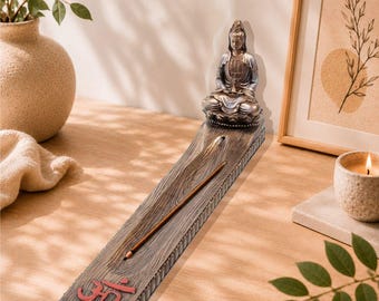 May include: A long, rectangular incense burner with a wood-grain texture, featuring a seated Buddha figure at one end and a red Om symbol at the other. A thin incense stick rests in the center groove of the burner, which has a lotus-petal border.