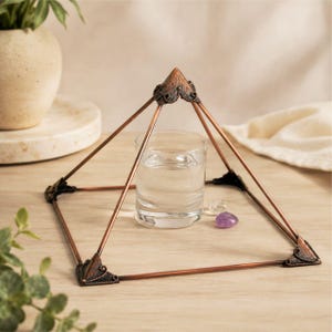 May include: A copper-coloured metal pyramid frame with ornate, dark-toned corner accents. The structure encloses a clear glass tumbler filled with water, accompanied by a small, translucent white stone and a smooth, purple amethyst gemstone.