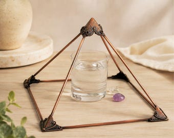May include: A copper-colored metal pyramid frame with ornate, dark-toned corner accents. The structure encloses a clear glass tumbler filled with water, accompanied by a small, translucent white stone and a smooth, purple amethyst gemstone.