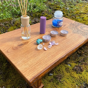 May include: A wooden altar table with a mandala design, featuring a diffuser bottle, candles, and crystals. The table has a natural wood finish and a rustic look.