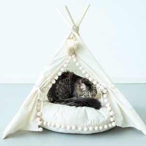 May include: A white canvas teepee pet bed with a fluffy white cushion and pom-pom trim. A tabby cat is resting inside the teepee.