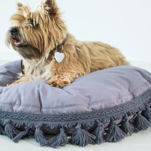 May include: A small, light brown dog with a white heart-shaped tag on its collar, lying on a round, gray dog bed with a gray tassel trim.