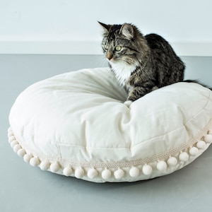 May include: A white round pet bed with a pom-pom trim. A gray and white cat is sitting on the bed.