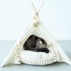 May include: A white canvas teepee pet bed with a fluffy white cushion and pom-pom trim. A tabby cat is resting inside the teepee.