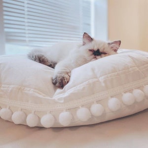 May include: A white fluffy cat with black markings on its face is sleeping on a white round pillow with a white pom-pom trim.