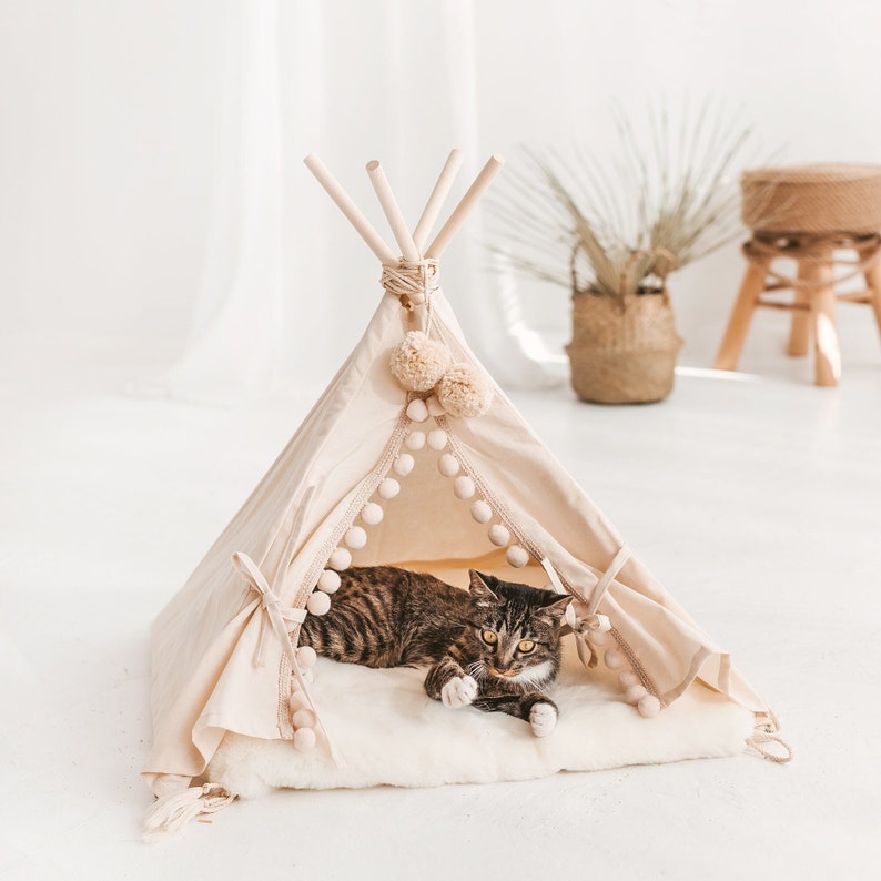 May include: A beige cat tent with a white faux fur lining and pom-pom trim. A tabby cat is lying inside the tent.