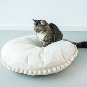 May include: A gray and white tabby cat sitting on a large, white, round pillow with a white pom-pom trim.