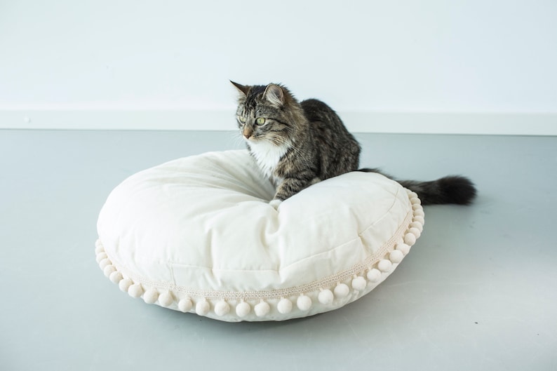 May include: A gray and white tabby cat sitting on a large, white, round pillow with a white pom-pom trim.