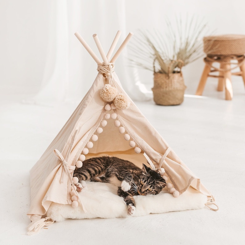 May include: A beige cat tent with a white faux fur cushion inside. The tent has a white pom-pom trim and a wooden frame. A tabby cat is lying inside the tent on the cushion.