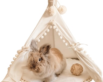 May include: A small, cream-colored teepee tent with a fluffy brown rabbit inside. The tent is decorated with white pom-poms and wooden poles. A ball of twine sits near the rabbit. The background is white.
