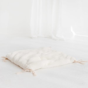 May include: A white faux fur cushion with a drawstring closure and tassels. The cushion is lying on a white surface.