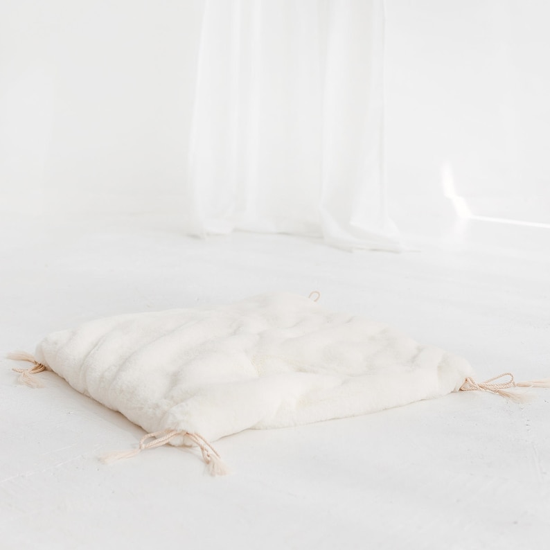 May include: A white faux fur cushion with a drawstring closure and tassels. The cushion is lying on a white surface.