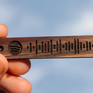 May include: A rectangular wooden keychain with a Spotify code engraved on it. The keychain is dark brown with a small Spotify logo on the left side. The code is made up of vertical lines of varying lengths.