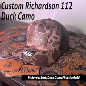Puede incluir: Gorra de béisbol Richardson 112 Duck Camo con parte trasera de malla marrón y parche de cuero marrón. El parche tiene el texto "SOUTHERN MAGNOLIA" y un logotipo. La gorra está rodeada de otros parches de cuero.