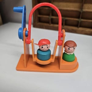 May include: A vintage plastic toy swing set featuring an orange base and frame with a blue hand crank. A small figure with a red cap sits in a blue swing, while a second figure with brown hair and a green base stands on the orange platform.
